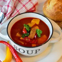 Soup in a bowl that is on a plate with bread and peppers next to it