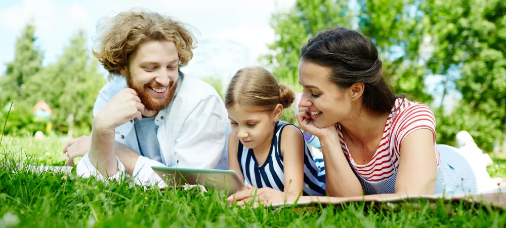 Family of three laying on the grass outside looking at a tablet